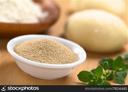 Active dry yeast in small bowl with fresh oregano on the side, flour and dough in the back (Selective Focus, Focus in the middle of the dry yeast) . Active Dry Yeast