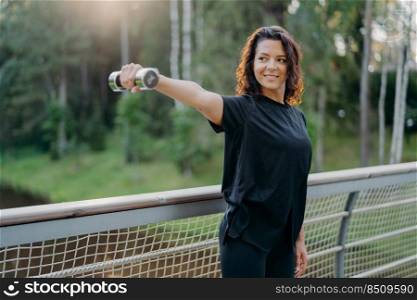 Active energized sportswoman stretches arms with dumbbell, poses on bridge over nature background with sunrise in morning, dressed in active wear, looks into distance, works on training biceps