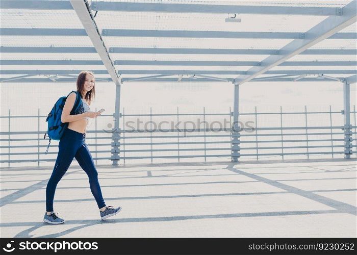 Active female fitness trainer dressed in sportsclothes, sneakers, carries rucksack, uses mobile phone for checking calories, listens music with earphones, uses free wifi connection. People and sport