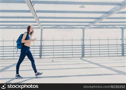 Active female fitness trainer dressed in sportsclothes, sneakers, carries rucksack, uses mobile phone for checking calories, listens music with earphones, uses free wifi connection. People and sport