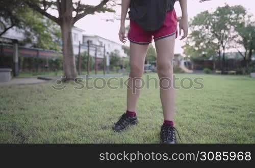 Active female stretching her legs standing on green grass lawn inside recreation park, lower body park, female holding legs doing warm up exercise before running, stretch quadriceps muscle flexibility