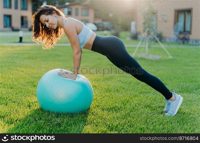 Active fitness woman does gymnastic outdoor with inflated fit ball, has active workout, makes physical exercises in open air, dressed in sport clothes trains on green grass. Active lifestyle concept
