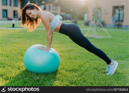 Active fitness woman does gymnastic outdoor with inflated fit ball, has active workout, makes physical exercises in open air, dressed in sport clothes trains on green grass. Active lifestyle concept
