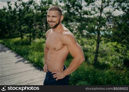 Active healthy lifestyle concept. Cheerful sporty man works out in nature, enjoys athlete exercising, keeps hands on hips, has muscular body, looks somewhere with cheerful smile does morning gymnastic