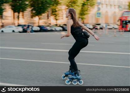 Active lifestyle and hobby concept. Sporty young woman does sport outdoors rides rollerblading dressed in sportsclothes enjoys fitness activities poses in urban place on road. Rollerskating.