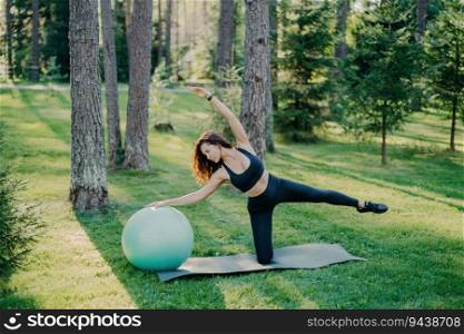 Active lifestyle concept: Sporty woman in black clothing leans on fitness ball, raises arms, poses on karemat in green park. Yoga training outdoors, showcasing an athletic body shape.