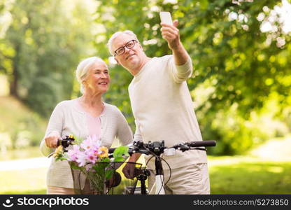 active old age, technology and lifestyle concept - happy senior couple walking with fixie bicycles taking selfie by smartphone at summer park. senior couple with bicycles taking selfie at park