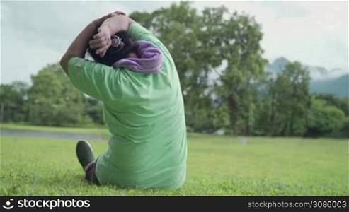 Active overweight senior woman stretching her arms shoulder sitting on grass lawn after exercise at the outdoor park, old age healthy life motivation after retirement, life insurance, middle aged