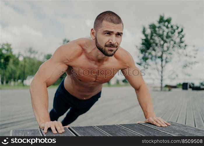 Activeμscular man does push up exercise poses shirt≤ss outdoor, exercises in park, has thick beard, stands in lower position. Motivated bodybuilder does workout®ularly. Ath≤te trains upper body