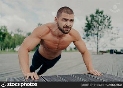 Activeμscular man does push up exercise poses shirt≤ss outdoor, exercises in park, has thick beard, stands in lower position. Motivated bodybuilder does workout®ularly. Ath≤te trains upper body