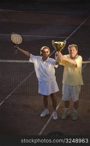 Active senior couple is posing on the tennis court with tennis racket and cup in hand. Outdoor, sunlight.
