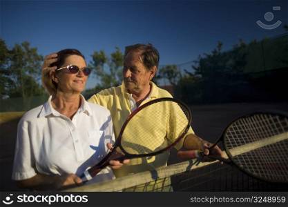 Active senior couple is posing on the tennis court with tennis racket in hand. Outdoor, sunlight.
