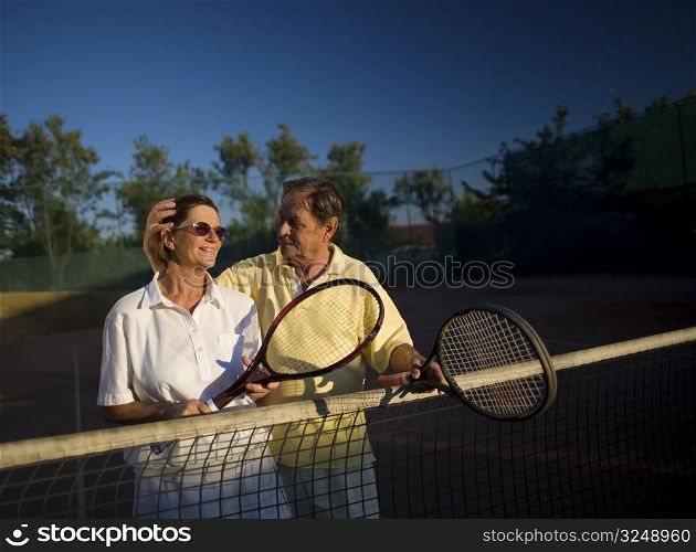 Active senior couple is posing on the tennis court with tennis racket in hand. Outdoor, sunlight.