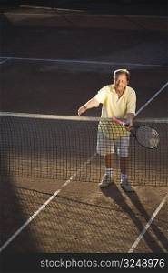 Active senior man in his 70s is offering his hand on the tennis court with tennis racket in other hand. Outdoor, sunlight.
