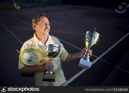 Active senior man in his 70s is posing on the tennis court with cups in hands. Outdoor, sunlight.