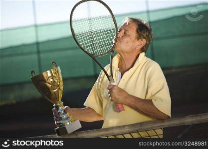 Active senior man in his 70s is very thankful to his tennis racklet while holding a cup in hand. Outdoor, sunlight.