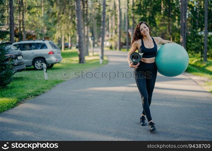 Active slim woman carries fitness ball and karemat, dressed in leggings, top, and sneakers. Walking near park or forest, with a gladful look and athletic body.