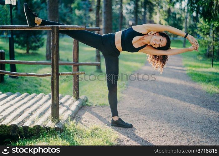 Active slim woman in sport clothes does pilates exercises and stretches outdoor, has glad expression, good flexibility, enjoys morning workout in fresh air. People, lifestyle, wellbeing concept