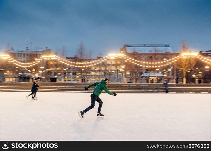 Active sorty male has fun in outdoor park on ice rink decorated with garlands, shows his talents of skating, makes fast movements on skates, being confident, enjoys winter outdoor activities