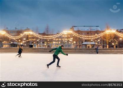 Active sorty male has fun in outdoor park on ice rink decorated with garlands, shows his talents of skating, makes fast movements on skates, being confident, enjoys winter outdoor activities