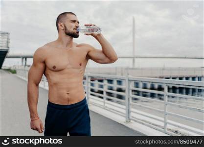 Active sportsman drinks bottle of water poses with naked torso concentrated into distance feels thirsty after running outdoors being full of energy, leads active lifestyle. Sport for better life