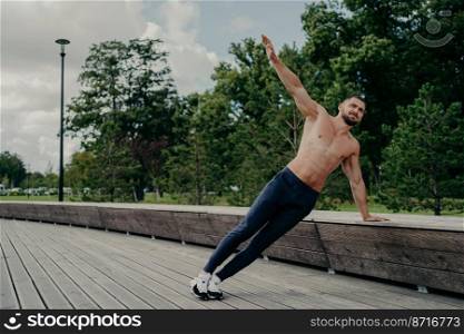 Active sportsman trains for strong healthy muscular body, poses outdoor in side plank pose, being trainer for himself, breathes fresh air. Abdominal workout concept. Determined guy trains patience