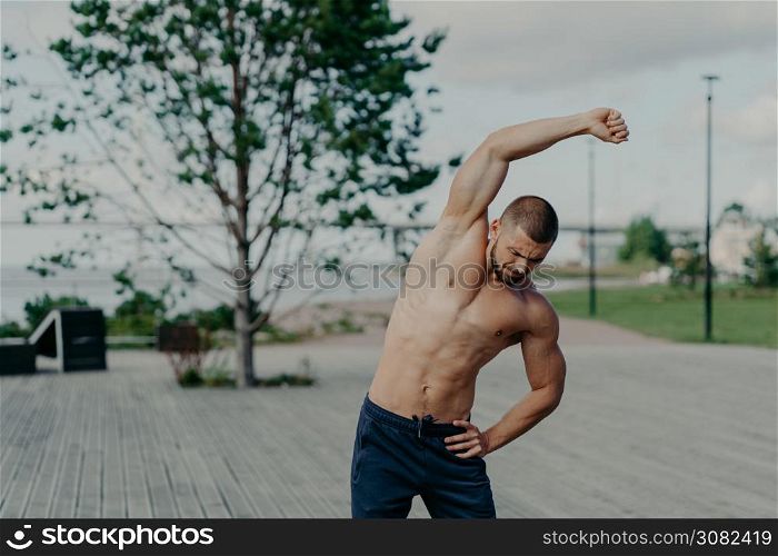 Active sportsman with naked torso and muscular body, does stretching exercises outdoor, shows good flexibility. Motivated athlete unshaven man warms up, prepares for workout, keeps muscles flexible.