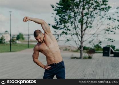 Active sportsman with naked torso and muscular body, does stretching exercises outdoor, shows good flexibility. Motivated athlete unshaven man warms up, prepares for workout, keeps muscles flexible.