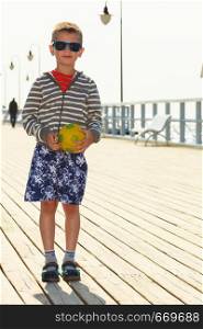 Active time on holidays concept. Young little boy holding yellow ball standing on pier outdoor. Kid child with toy on summer holiday time.. Boy with ball on pier.