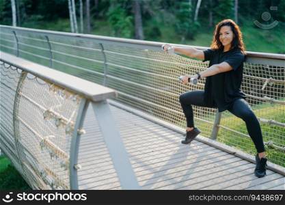 Active woman does squats with dumbbells at a bridge, wearing active wear, training biceps with sport equipment. Expresses joy in sporty lifestyle concept.