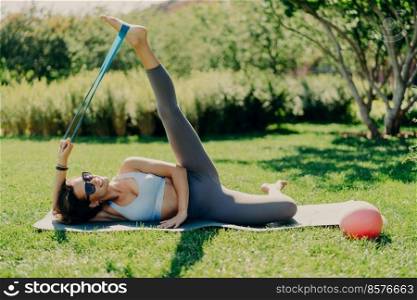 Active woman does workout outside. Slim brunette woman stretches legs with sports gum dressed in activewear lying on fitness mat against green nature background has good flexibility. Exercising