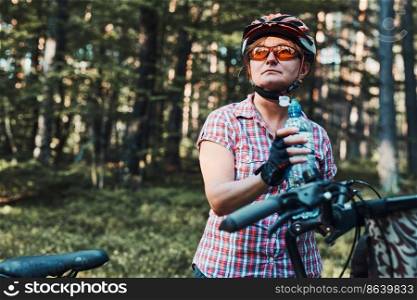 Active woman spending free summer vacation time on a bicycle trip in a forest. Woman wearing bicycle helmet and gloves holding bike with basket and drinking a water from bottle. Active woman spending vacation summer time on a bicycle trip in a forest