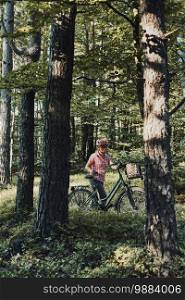 Active woman spending free summer vacation time on a bicycle trip in a forest. Woman wearing bicycle helmet and gloves holding bike with basket standing behind the trees. Active woman spending free summer vacation time on a bicycle trip in a forest