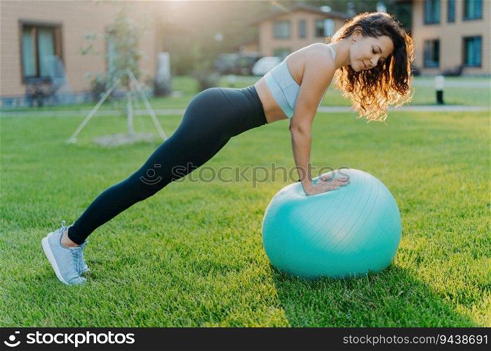 Active woman trains outdoors, does gymnastics with fit ball, wears sport clothes, exercises on green grass. Active lifestyle.