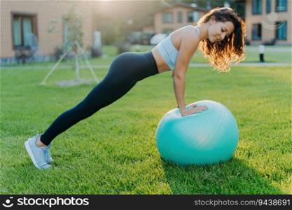 Active woman trains outdoors, does gymnastics with fit ball, wears sport clothes, exercises on green grass. Active lifestyle.
