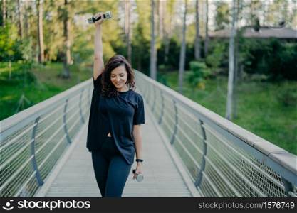 Active young brunette sportswoman exercises with dumbbells, does fitness exercises outdoor, dressed in active wear, breathes fresh air and works on muscles. Women, strength and bodybuilding concept