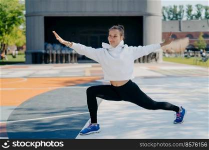 Active young woman dedicated to fitness does stretching exercises spreads hands dressed in active wear stretches legs and arms poses outdoo. People regular sport activities and lifestyle concept