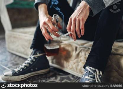 Addicted male person sitting on the mattress with cigarette and bottle of alcohol in hands, grunge room interior on background. Addiction concept. Addicted male person with cigarette and alcohol