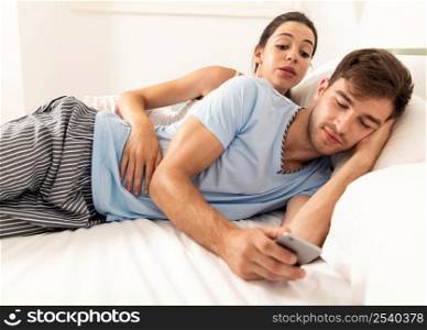 Addicted young man on bed texting while woman looks angry