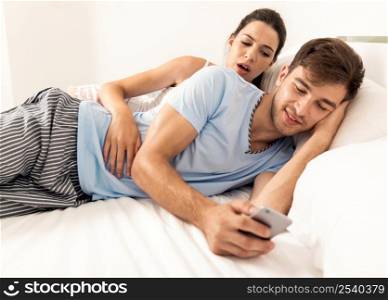 Addicted young man on bed texting while woman looks angry