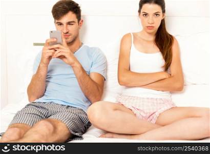 Addicted young man on bed with a cellphone while the woman looks angry