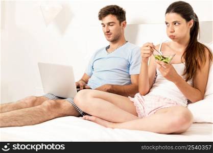 Addicted young man on bed with a laptop while the woman looks angry
