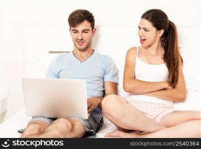 Addicted young man on bed with a laptop while the woman looks angry