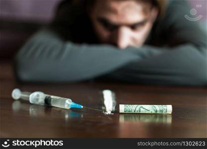 Addiction man look at the table with syringe, dollars and cocain trace. Abuse of narcotics leads to a depression.