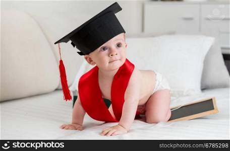 Adorable 10 months old baby boy in graduation cap crawling on bed