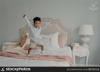 Adorable african cute kid jumping on bed covered with bedspreads in light pink and white tones next to l&on bedside table, active mixed race child playing in parents bedroom during leisure time. Adorable cute kid jumping cheerfully on bed and having fun at home