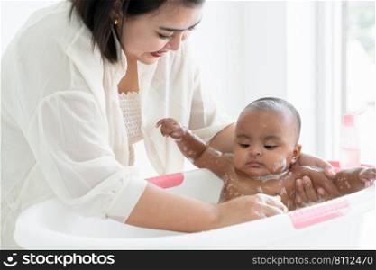 Adorable African newborn baby bathing in bathtub. Asian young mother use sponge with shower gel made soap bubbles to wash her little daughter in warm water. Newborn baby cleanliness care concept