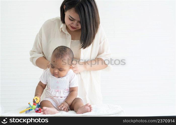 Adorable African newborn girl sitting and holding toy in hand while young Asian mother dressing changing her infant clothes after bath at home. White background. Bonding in mixed race family