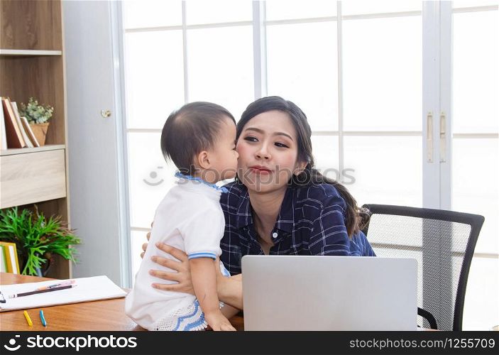 Adorable Asian baby girl kiss beautiful mom with love spending time together at home office small business, single mother working on laptop technology computer, parenthood working and nursing baby