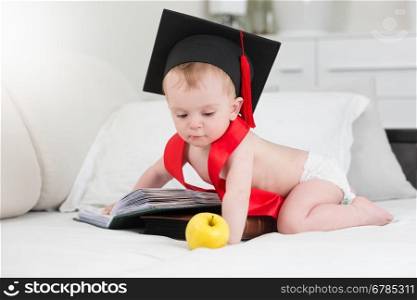Adorable baby boy in graduation cap posing with apple and big book